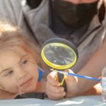 young girl holding magnifying glass looking at small praying mantis on paintbrush