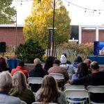 people sitting outside in chairs with speaker at podium and tv screen
