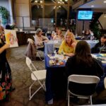 group of people sitting at tables watching woman with finished art project