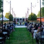 people sitting in chairs alongside an aisle lined with string lights in front of a person at a podium