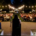 person standing at a podium in front of a crowd of people sitting down outside surrounded by string lights