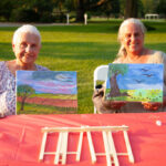 two women smiling holding up their painting on canvas